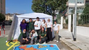 La foto ritrae nove persone davanti a una tenda bianca, in un contesto urbano. Alcuni indossano magliette verdi o bianche e hanno segni di colore sulle braccia. Davanti a loro è steso un grande telo colorato con forme e disegni, che richiama un’attività creativa o partecipativa svolta all’aperto.