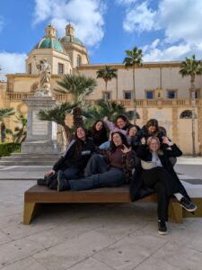 7 persone sedute su una panchina in una piazza, con un grande edificio storico alle loro spalle, c