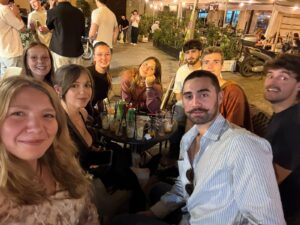  nove persone sedute attorno a un tavolino rotondo in un bar all’aperto, durante la sera. Sul tavolo ci sono bicchieri, lattine e cannucce. Intorno si vedono altre persone, piante e luci decorative, creando un’atmosfera vivace e sociale.