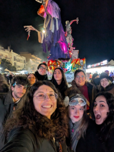 L’immagine mostra un gruppo di persone che posa durante un evento notturno all’aperto. Alle loro spalle c’è una grande installazione artistica illuminata con colori vivaci, raffigurante una figura scenografica con tessuti colorati e decorazioni.